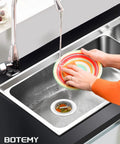 Person rinsing a colorful plate over a kitchen sink with 304 Stainless Steel Kitchen Sink Drain Strainer Assembly Kit For 3.5 Inch Drains Built To Last Heavy Duty Rustproof And Leakproof Basket Stopper With Universal Fit For Standard American Sinks Brushed Finish visible.