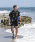 Man wearing Glide Front Adjustable Scuba BCD With Balanced Power Inflator For Recreational Diving All Day Comfort with Y Fit Shoulders Neoprene Neck Pad And Adjustable Cummerbund XL Blue, walking on beach.